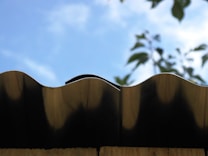 A close-up view of a wavy metal roof edge against a bright blue sky. In the background, blurred green leaves partially visible, adding a natural contrast.