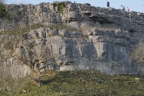 A rugged rock cliff with layers of stone formations. There is a leafless tree at the base of the cliff, set against a backdrop of green grass. Several people can be seen walking along the top, suggesting a hiking or outdoor exploration setting.