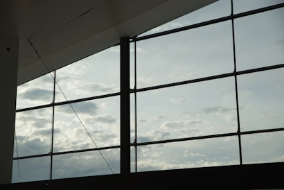 Large glass windows reveal a cloudy sky, with soft light filtering through. The metal frames create a geometric pattern against the sky, capturing a modern architectural element.