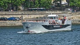 A passenger boat named 'Paris Montmartre' navigates a river with people on board enjoying the ride. The scene includes a stone embankment with trees and parked cars in the background, indicating a city environment.