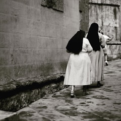 Two women dressed in long, dark veils and light robes walk along a narrow cobblestone street beside a textured stone wall. The setting has an old-world feel, suggesting a historical or religious context.