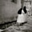 Two women dressed in long, dark veils and light robes walk along a narrow cobblestone street beside a textured stone wall. The setting has an old-world feel, suggesting a historical or religious context.
