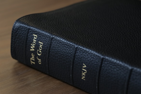 A black leather-bound book is displayed on its side with gold lettering along the spine indicating it is titled 'The Word of God' and marked as 'NKJV'. The surface on which the book rests appears smooth and wooden.