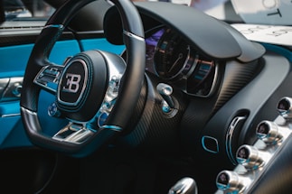 A close-up view of a luxury car's interior, featuring a steering wheel with a prominent logo, a sleek dashboard with a speedometer, and various metallic control knobs. The interior design includes high-quality materials, such as leather and carbon fiber.