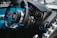 A close-up view of a luxury car's interior, featuring a steering wheel with a prominent logo, a sleek dashboard with a speedometer, and various metallic control knobs. The interior design includes high-quality materials, such as leather and carbon fiber.