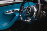 A luxurious car interior featuring a steering wheel with a prominent logo in the center. The interior is adorned with sleek metallic and black components, contrasted by vibrant blue accents on the door and dashboard. The image highlights the sophistication and elegance of the vehicle's design.