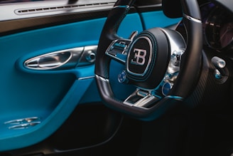 A luxurious car interior featuring a steering wheel with a prominent logo in the center. The interior is adorned with sleek metallic and black components, contrasted by vibrant blue accents on the door and dashboard. The image highlights the sophistication and elegance of the vehicle's design.