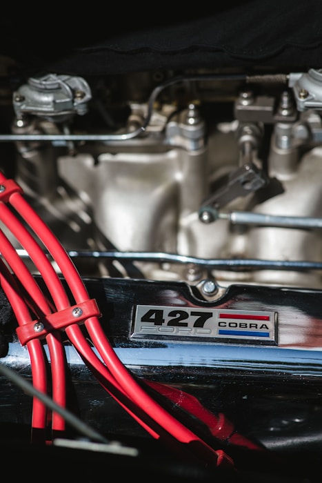 An engine compartment showcasing chrome and metallic components combined with a prominent label reading '427 Ford Cobra'. Bright red cables are neatly bundled in the foreground, adding a vivid contrast to the otherwise metallic hues of the engine parts.