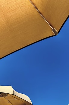 A clear blue sky is partially covered by two large sunshades. The sunshades are tan in color, with one occupying the top right corner and the other positioned at the bottom left. The fabric texture of the sunshades is clearly visible, giving a sense of relaxation and protection from the sun.