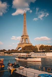 A scenic view of the Eiffel Tower surrounded by lush greenery and the Seine River in the foreground. Multiple boats are visible on the water, with people leisurely enjoying the river cruise. The sky is clear with a few scattered clouds, and the lighting suggests a late afternoon or early evening setting.