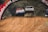 Two cars are airborne on a dirt track under a large arch with a red and white advertisement. They appear to be in a competitive race surrounded by a wooded area. The track is uneven and dusty, indicating a challenging racing environment.