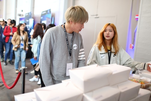 Several people are in a line at what appears to be an event or conference. Two individuals in the foreground are engaged in conversation near a table with stacked white boxes and packaged goods. The atmosphere is casual, with attendees wearing lanyards.