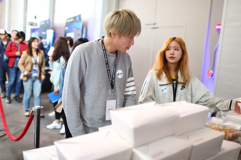 Several people are in a line at what appears to be an event or conference. Two individuals in the foreground are engaged in conversation near a table with stacked white boxes and packaged goods. The atmosphere is casual, with attendees wearing lanyards.