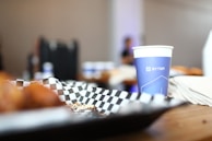 A blue paper cup with a logo and geometric design stands on a wooden table surrounded by blurred background objects. A black and white checkered paper is nearby, with some crumbs visible on it. The background is softly out of focus, contributing to a casual and relaxed setting.
