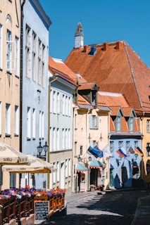 A quaint cobblestone street lined with charming buildings featuring colorful facades, predominantly in pastel shades. The buildings display a mix of pale blues, beiges, and reds with red-tiled roofs. Vibrant flowers adorn the edge of an outdoor seating area, next to which stand several flags of different countries. A clear blue sky serves as a backdrop, adding to the serene atmosphere.