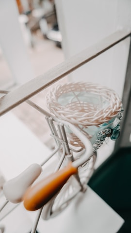 A decorative, miniature bicycle with a woven basket attached is placed near a window. The bicycle features a white seat and orange handlebars, with detailed metal work and lace decorations near the basket.