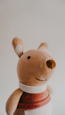 A plush toy resembling an animal, possibly a kangaroo or bear, with a soft fabric texture. It features a light brown color with darker brown on the nose and an embroidered smile. It wears a maroon and white outfit, and its ears have white inner patches.
