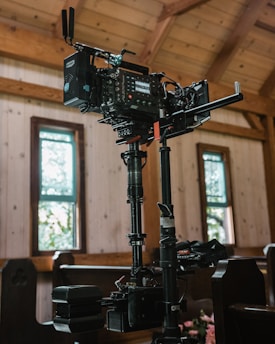 A professional camera setup is positioned indoors, likely in a wooden structure with natural light coming through vertical windows. The camera is mounted on a stabilizing rig indicating preparations for filming.
