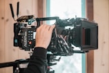 A professional cinema camera is being adjusted by a person. The camera has multiple attachments and is mounted on a support system, with a lens and matte box visible. A wooden wall and a window in the background suggest an indoor setting with natural light.