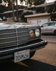 A classic car with a vintage design is parked on a street in front of a residential area. The car's grille and headlight are prominently featured, along with a California license plate. Another modern car is partially visible in the background.