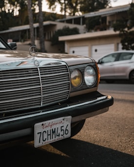 A classic car with a vintage design is parked on a street in front of a residential area. The car's grille and headlight are prominently featured, along with a California license plate. Another modern car is partially visible in the background.