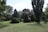 A serene landscape featuring an assortment of evergreen trees and well-maintained grass. The scene includes a variety of tree species with different shapes and sizes, set under a clear blue sky with scattered clouds.