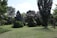 A serene landscape featuring an assortment of evergreen trees and well-maintained grass. The scene includes a variety of tree species with different shapes and sizes, set under a clear blue sky with scattered clouds.