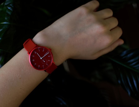 A wrist wearing a bright red watch with a simple, minimalist design. The watch has clear numerals and white hands against the red background. The background consists of dark green leaves, creating a natural and elegant contrast with the watch.