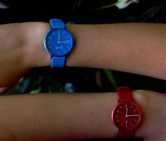 Two wrists are adorned with watches, one blue and one red. The watches have minimalist designs with no numerals and contrasting second hands. The lighting casts a soft shadow on the skin, emphasizing the simplicity and elegance of the timepieces.