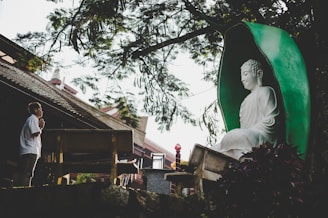 A serene scene with a large white Buddha statue encased in a green alcove surrounded by trees. A person stands nearby in a prayer position, showing reverence. The setting includes elements of traditional architecture and a peaceful natural environment.