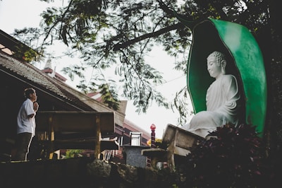 A serene scene with a large white Buddha statue encased in a green alcove surrounded by trees. A person stands nearby in a prayer position, showing reverence. The setting includes elements of traditional architecture and a peaceful natural environment.