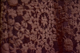 A close-up view of a textured fabric with a delicate lace pattern. The lace is intricately woven with floral designs, creating a detailed and elegant appearance.