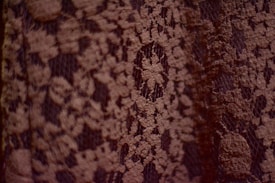 A close-up view of a textured fabric with a delicate lace pattern. The lace is intricately woven with floral designs, creating a detailed and elegant appearance.
