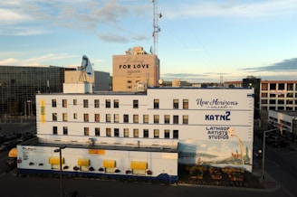 A multi-story building adorned with various signs, including 'New Horizons Gallery,' 'Lathrop Artists Studios,' and 'KATN 2.' The facade is painted with a mural depicting a scenic landscape. The surrounding area includes a few other buildings, visible under a clear sky with some clouds. An advertisement with the phrase 'Looking for Love Again' is displayed on an adjacent building. There are parked cars and a road in the foreground.