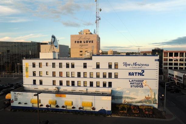 A multi-story building adorned with various signs, including 'New Horizons Gallery,' 'Lathrop Artists Studios,' and 'KATN 2.' The facade is painted with a mural depicting a scenic landscape. The surrounding area includes a few other buildings, visible under a clear sky with some clouds. An advertisement with the phrase 'Looking for Love Again' is displayed on an adjacent building. There are parked cars and a road in the foreground.
