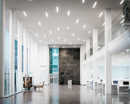 A spacious, modern interior with tall white columns and high ceilings. The floor is polished and reflective. On the left side, there is a comfortable seating area and a glass display, while the right side features modern desks with computer stations. The walls are a mix of white and dark gray panels, and there is ample natural light from large windows.