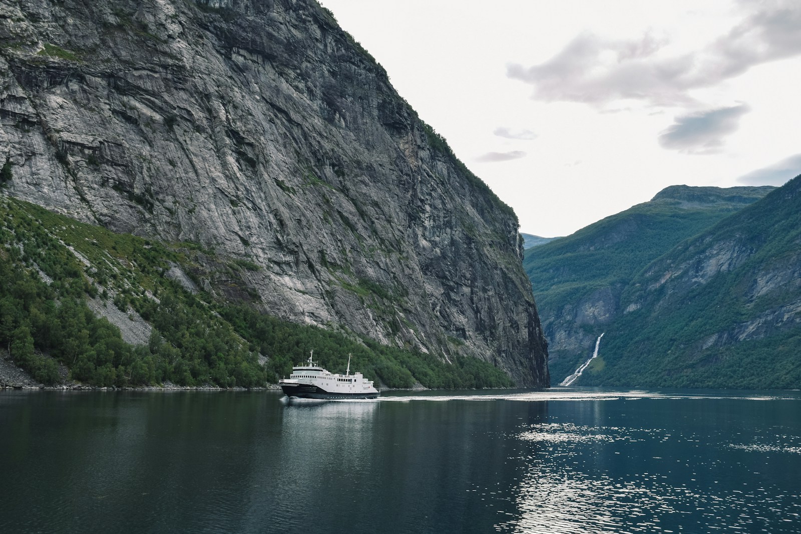 A white passenger ship sailing a narrow fjord at the foot of high cliffs, with a thin waterfall down the rock