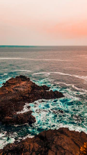 A sweeping view of a rugged coastline at sunset, waves crashing against jagged cliffs under a deep blue sky.