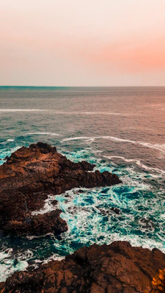 A rugged coastline at dusk with teal and navy waves crashing against weathered rocks, accented by a splash of red in the sky.