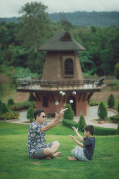 A man and a child sit on a grassy field, engaged in a playful juggling activity with multiple small balls mid-air. In the background, a rustic windmill structure is surrounded by lush greenery and ornamental plants, creating a serene and picturesque scene.