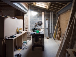 A cluttered workshop with a variety of wood pieces and tools scattered around. A miter saw is prominently placed on a worktable in the middle. There are wooden planks leaning against the walls and various construction materials and equipment like a tape measure and boxes are visible. The space is illuminated by natural light coming through a skylight.