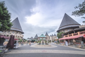 A courtyard surrounded by medieval castle-like buildings with conical rooftops. The center contains a decorative fountain, and the area is paved with stone tiles. There are a few people strolling and some market stalls on the sides. The sky is overcast, giving a serene atmosphere.