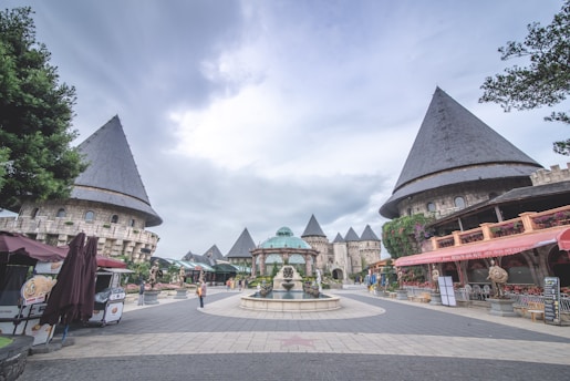 A courtyard surrounded by medieval castle-like buildings with conical rooftops. The center contains a decorative fountain, and the area is paved with stone tiles. There are a few people strolling and some market stalls on the sides. The sky is overcast, giving a serene atmosphere.