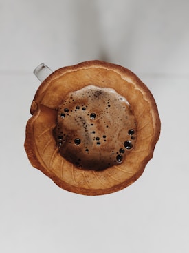 A top-down view of a coffee filter placed on a dripper, filled with freshly brewed coffee. The filter is visibly wet and dark in color with the texture of coffee grounds. The coffee surface has visible bubbles and a dark, rich appearance. The dripper handle is partially visible on the side.