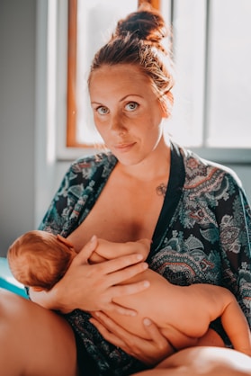 A woman with auburn hair in a bun sits in a softly lit room, wearing a patterned robe and breastfeeding a baby.