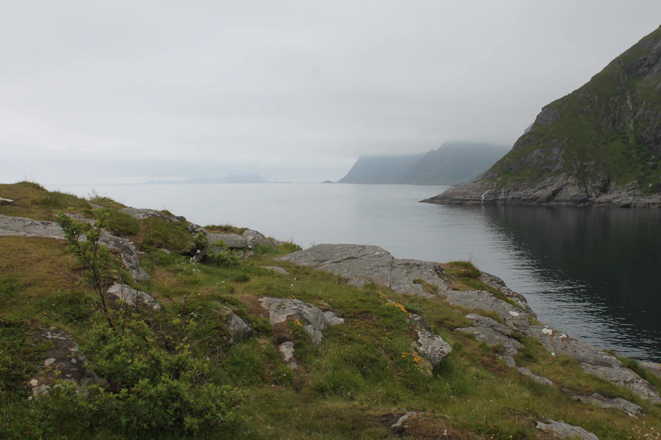 The image depicts a serene landscape with a rocky cliff overlooking a body of water, possibly an ocean or sea. The cliff is covered in green grass and small plants, adding a touch of life to the rugged terrain. In the distance, there are several islands visible on the horizon, their white and gray hues contrasting against the overcast sky. The perspective of the image suggests it was taken from a higher vantage point, providing an expansive view of the landscape below.