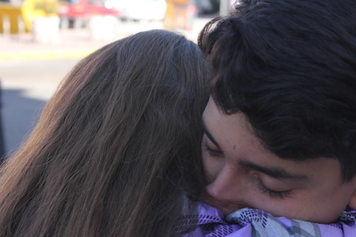 Two people are embracing each other closely, conveying a sense of warmth and affection. The background is slightly blurry, focusing attention on the emotional moment.
