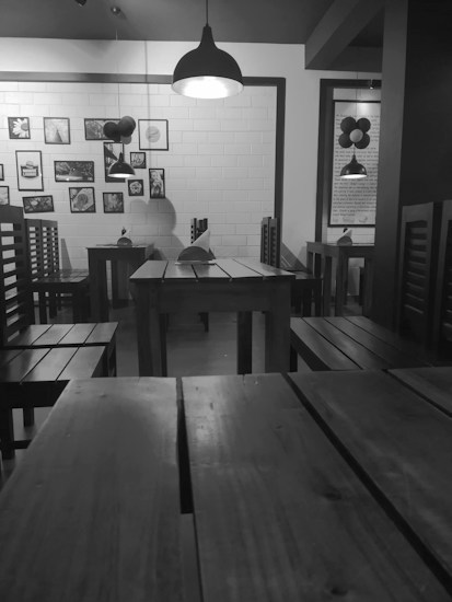 A dimly lit indoor dining area features wooden tables and chairs with a modern, minimalist design. Pendant lights hang from the ceiling, casting soft light on the tables below. Various framed artworks and photographs hang on a white-tiled wall. Balloons are tied to one of the chairs, adding a touch of festivity.