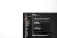 A computer monitor on the right side of the image displays a text editor with code written in a dark theme. The text editor interface includes a file explorer on the left showing a list of project files, while the right side presents code written in JavaScript, involving elements like functions and error handling. The screen is set against a stark white background occupying the left part of the image.