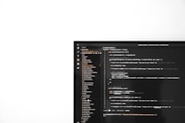 A computer monitor on the right side of the image displays a text editor with code written in a dark theme. The text editor interface includes a file explorer on the left showing a list of project files, while the right side presents code written in JavaScript, involving elements like functions and error handling. The screen is set against a stark white background occupying the left part of the image.